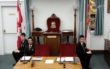 An image of the clerk's table and the speaker's chair in the legislative chamber, with pages seated on the dais