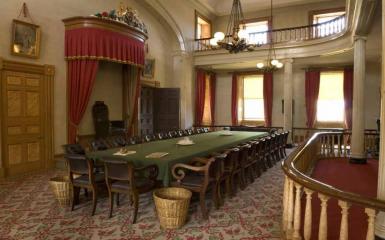 An image of the Confederation Chamber in Province House, with a chair on a dais and a long table covered with a green cloth